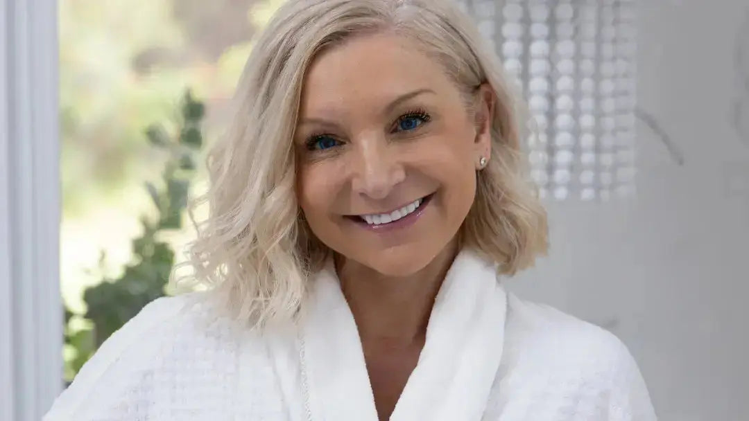 Woman in a white robe holding a STMNA Bioactives product - Healthspan and a package, with a blurred indoor background.