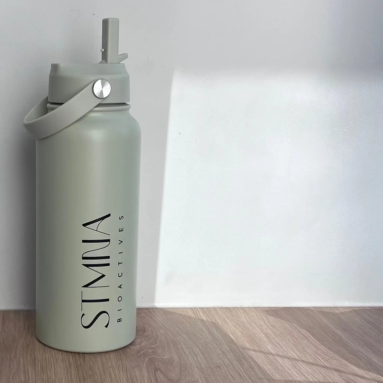 A matte sage green stainless steel water bottle with a carry loop and flip spout lid, featuring the brand name 'STMNA Bioactives' printed vertically on the front, set on a light wood floor against a white wall.