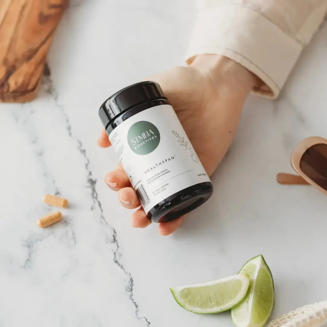 Hand holding a supplement container labeled STMNA Bioactives on a marble surface with lime wedges.