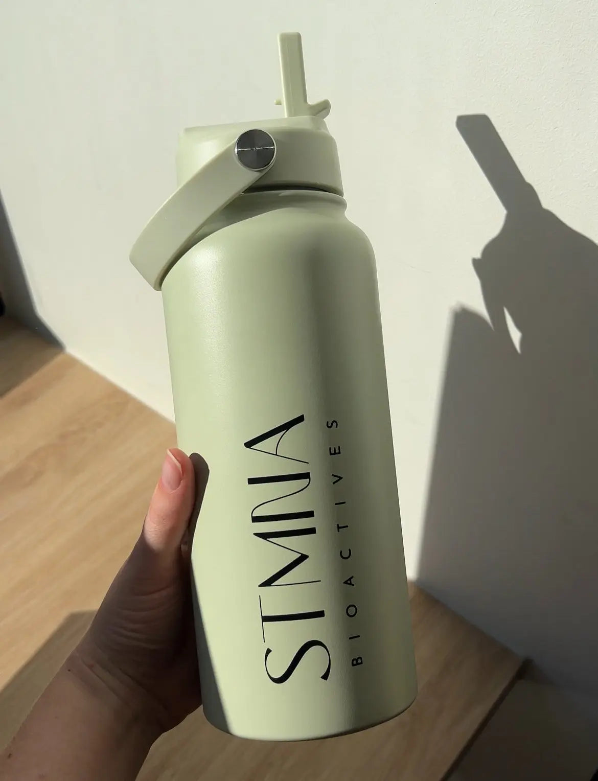 A hand holding a matte sage green water bottle with a flip-top lid and black vertical branding that reads 'STMNA BIOACTIVES'. The photo is taken in a real indoor setting with a wooden table and a wall, casting a strong shadow.