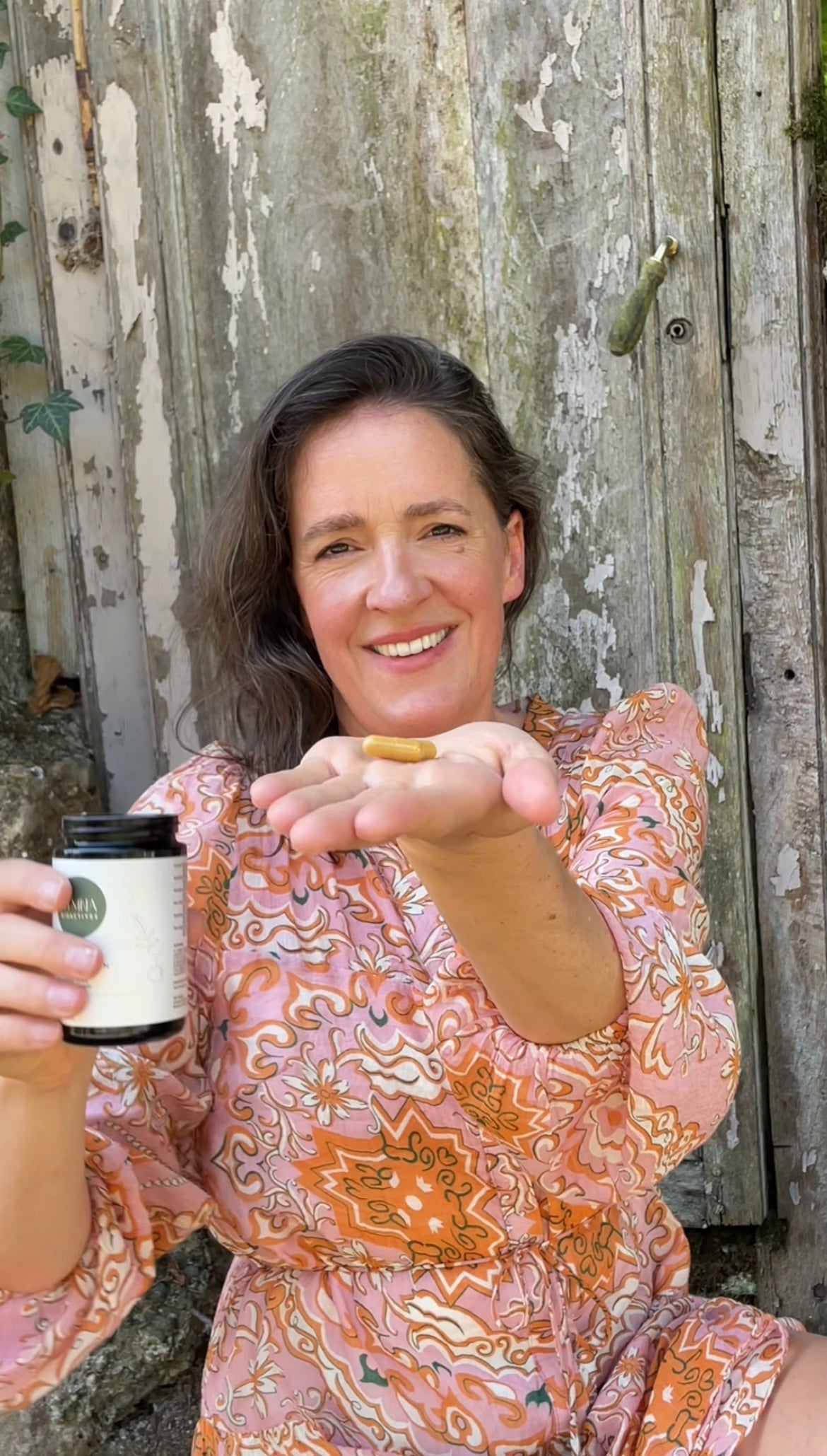 Woman holding a STMNA Bioactives Jar and a daily capsule in front of a wooden fence