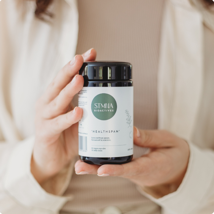 Person holding a supplement container labeled 'STMNA Bioactives Healthspan' against a neutral background