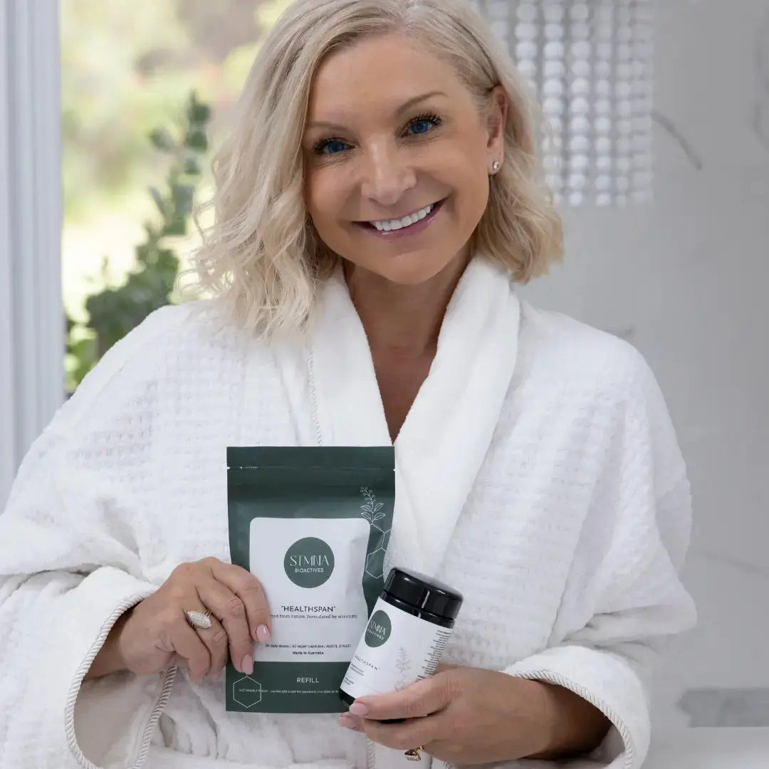 Woman in a white robe holding a STMNA Bioactives product - Healthspan and a package, with a blurred indoor background.
