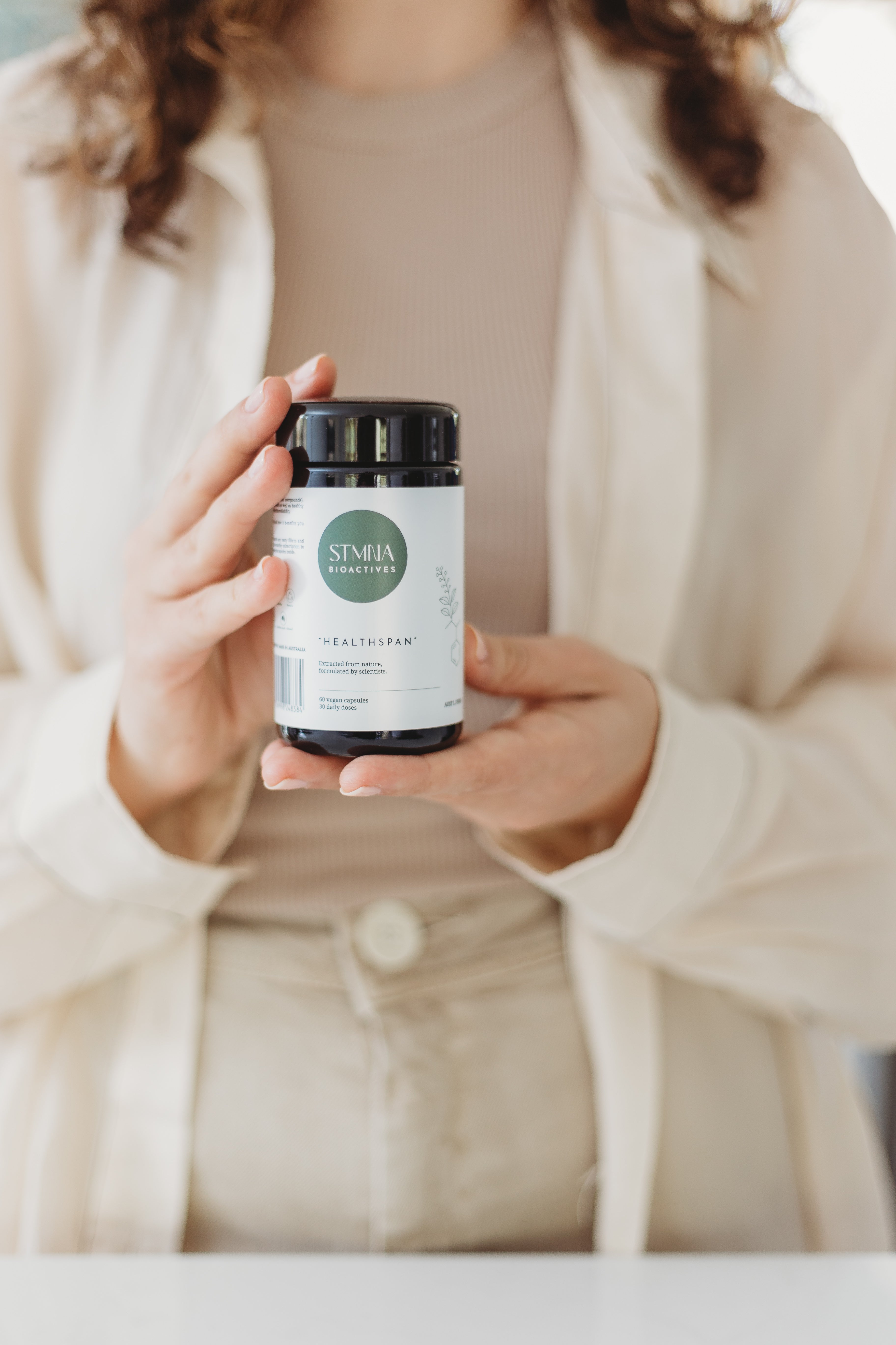 Person holding a supplement container labeled 'STMNA' in a neutral setting